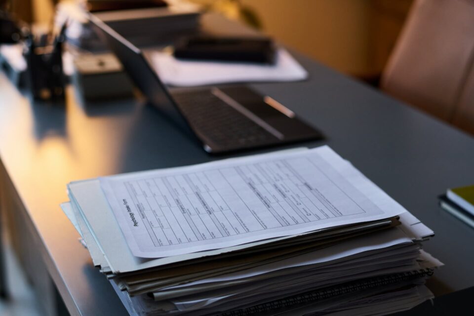 Stack of printed documents in a dark office with a laptop visible in the background