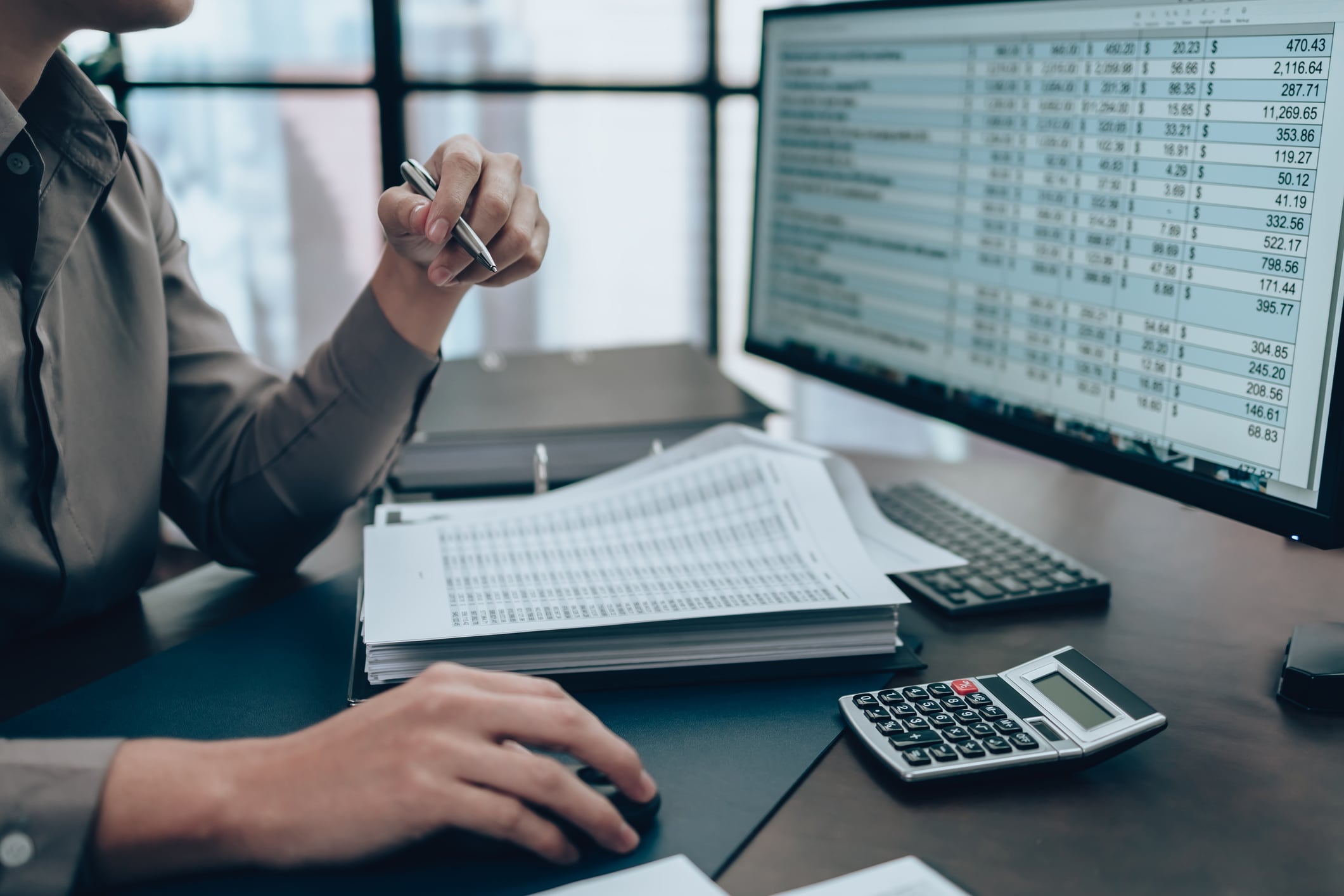 Person pointing at a spreadsheet on a monitor while using a calculator and keyboard