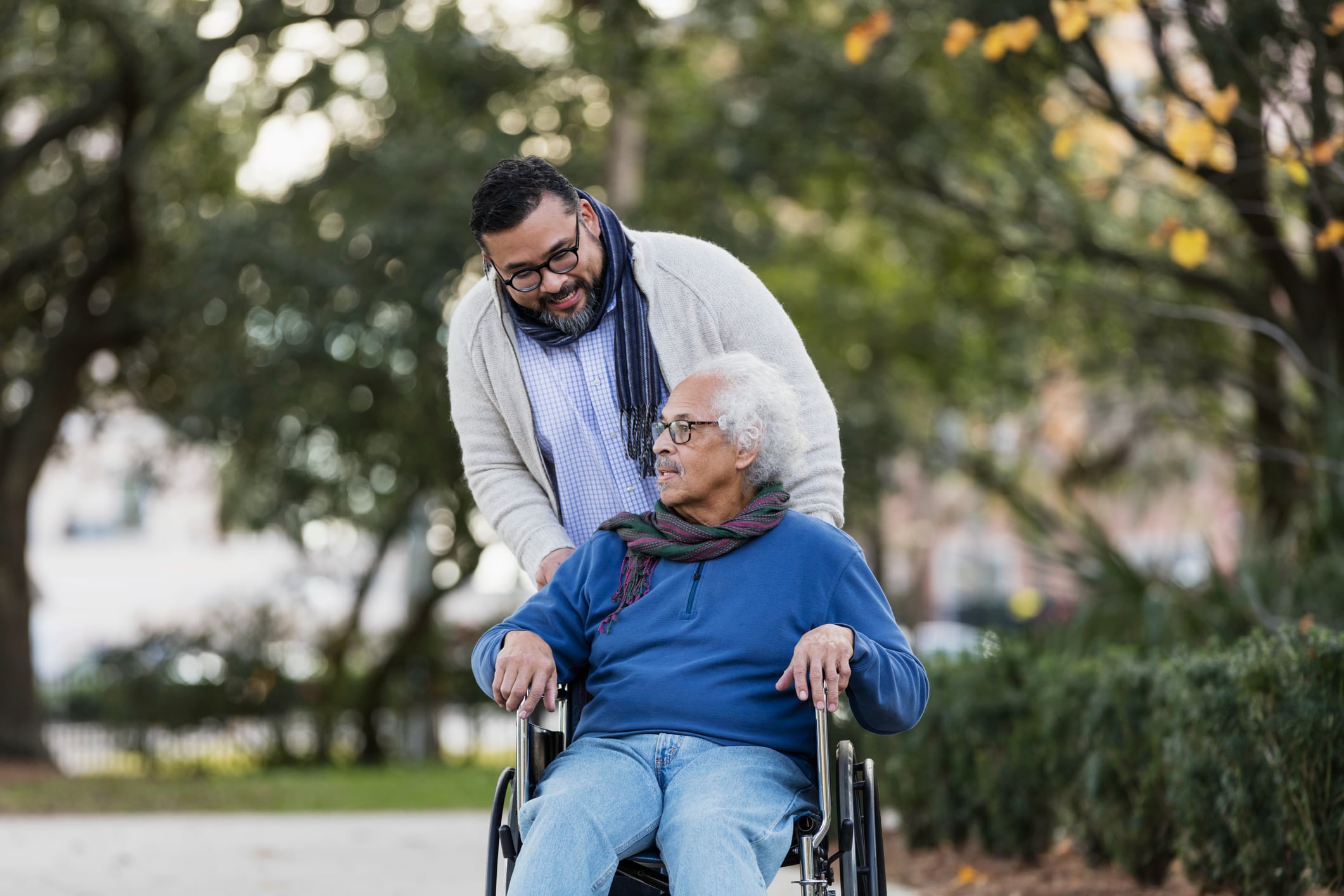 Man pushing an elderly woman in a wheelchair through a tree-lined park