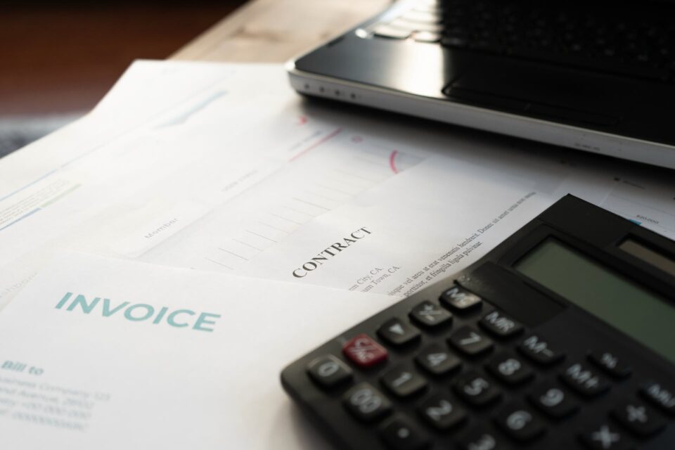 Invoice document next to a calculator on a desk with a laptop and contract visible