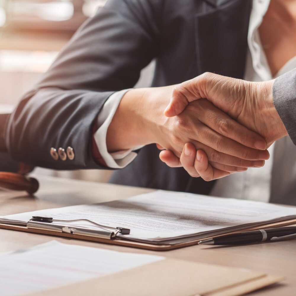 Two people in business attire shaking hands over a desk with a contract