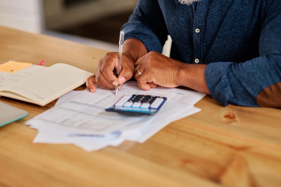 Older person seated at a table with documents and a calculator, writing
